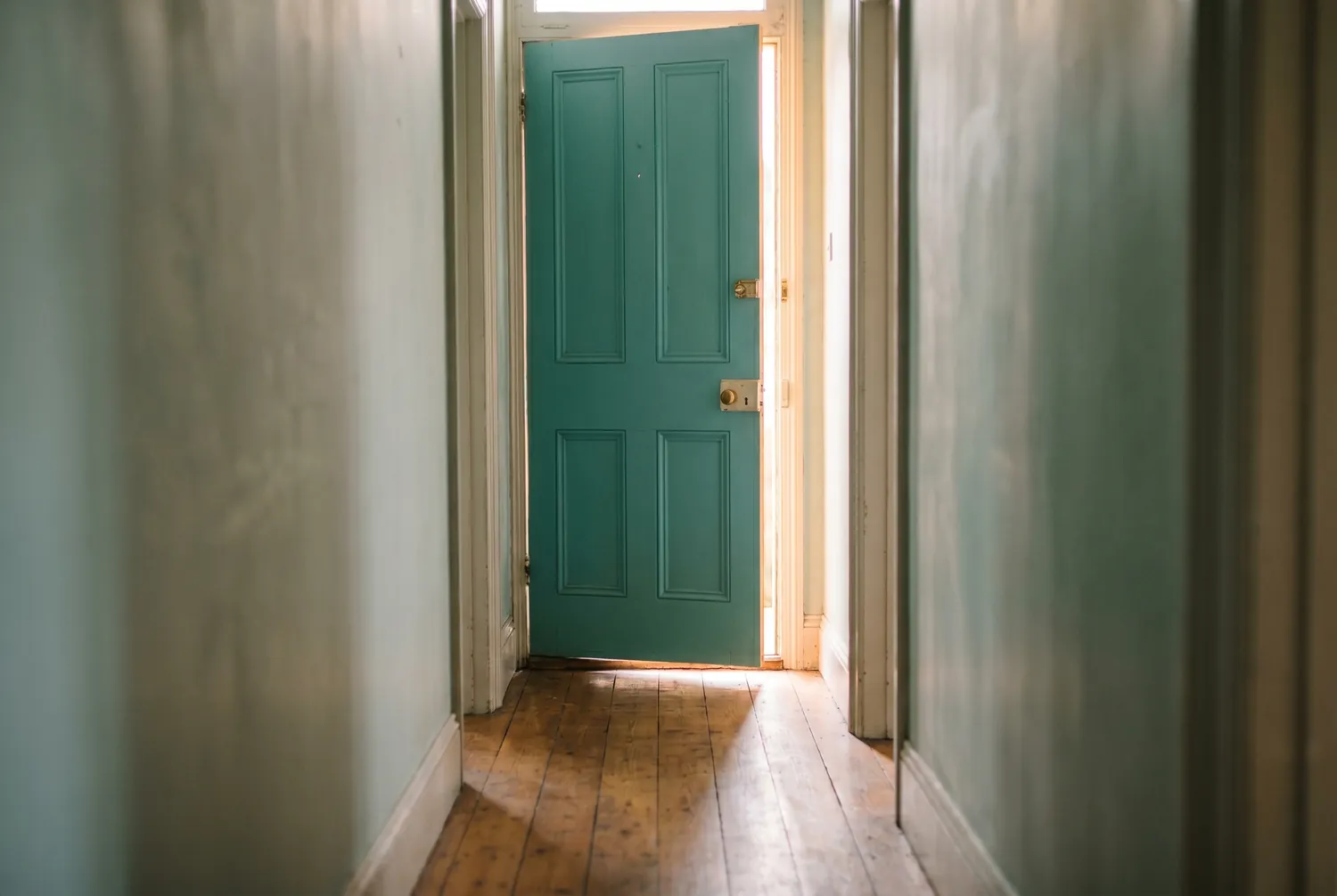 Morning light spilling through a teal front door standing slightly ajar at the end of a hallway