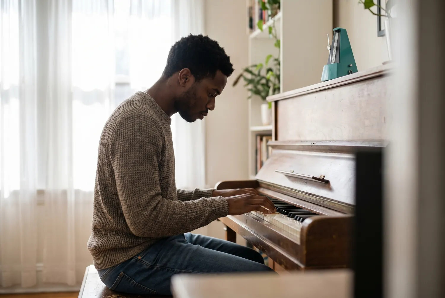 Young man practicing scales at an upright piano in morning light with a single teal metronome on the piano top