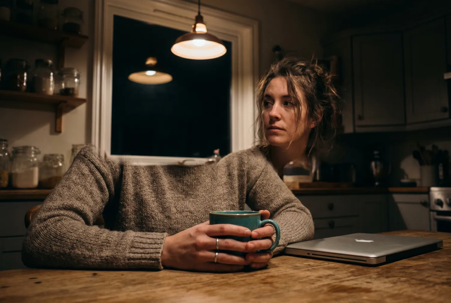 Woman sitting at a kitchen table late at night holding a single teal mug, gaze drifting toward the dark window Woman sitting at a kitchen table late at night holding a single teal mug, gaze drifting toward the dark window