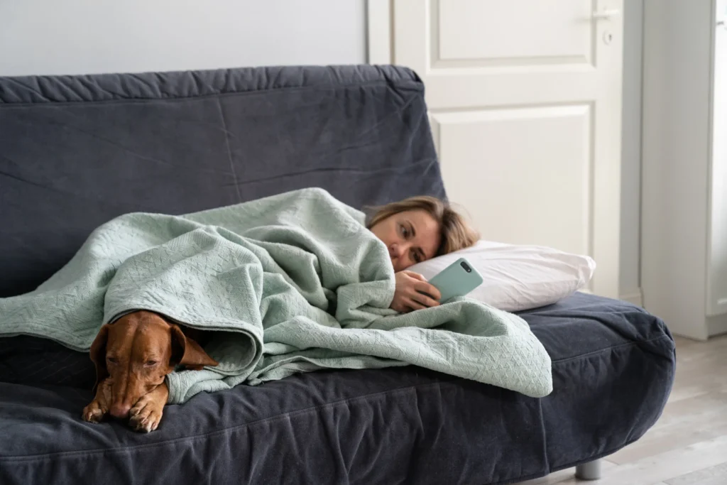 woman staring at her phone while laying on couch with a dog