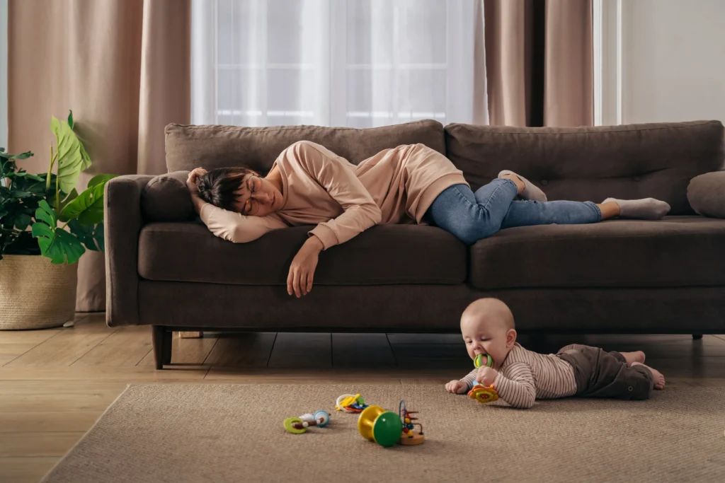 mother falling asleep on couch while her baby is on the floor in front of her