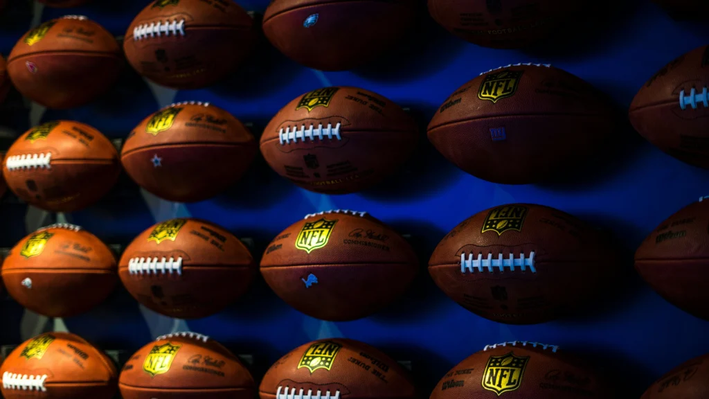 Wall display of multiple signed Wilson NFL footballs with team logos and commissioner signatures under blue lighting, illustrating legacy and mental pressure in elite football like the Super Bowl